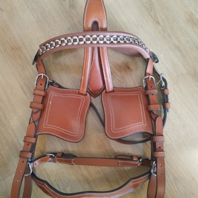 Arnês de couro castanho com costuras brancas e fivelas metálicas sobre piso madeira