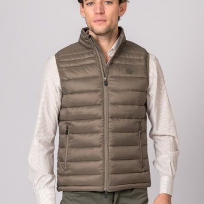Homem a usar colete acolchoado castanho com fecho frontal e bolsos, camisa bege e calças verdes
