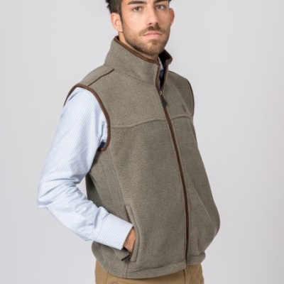 Homem com colete cinza de tecido polar e fecho castanho, camisa azul clara e calças castanhas sobre fundo cinza claro.