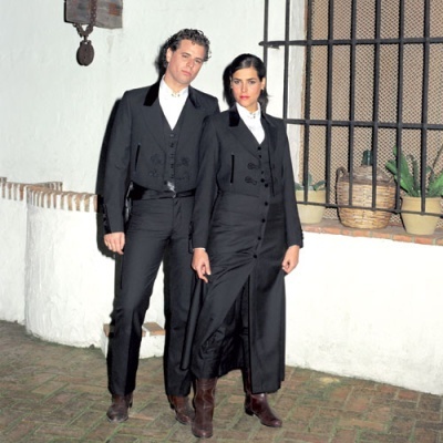 Dois modelos com trajes tradicionais pretos em ambiente exterior com plantas.