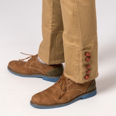 Sapatos masculinos castanhos de camurça com sola azul junto a calças bege com botões vermelhos na barra