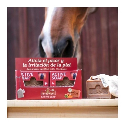 Sabão artesanal ACTIVE SOAP em embalagem vermelha com texto em espanhol e sabão com toalha branca em superfície de madeira com cavalo ao fundo