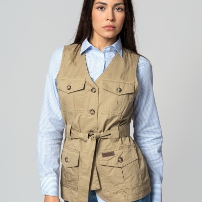 Mulher com colete bege de bolsos e cinto, camisa azul clara às riscas e jeans azuis.