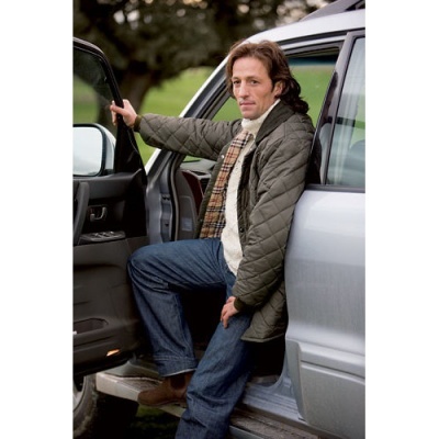 Homem sentado na soleira de carro prateado com casaco acolchoado cinza e jeans