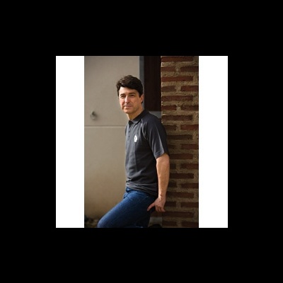 Homem com camisa preta e calças azuis encostado numa parede de tijolos vermelhos