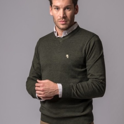 Homem a vestir camisola verde escuro de malha com gola em V sobre camisa xadrez clara e calças castanhas.