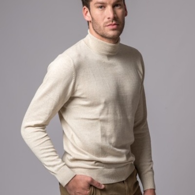 Homem com camisola de gola alta bege e calças castanhas num fundo cinza