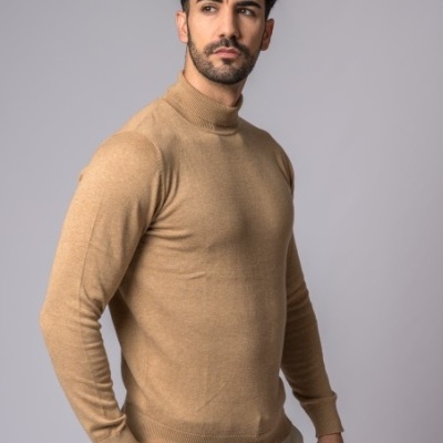 Homem com camisola de gola alta castanha clara e calças bege claras, fundo cinza liso.