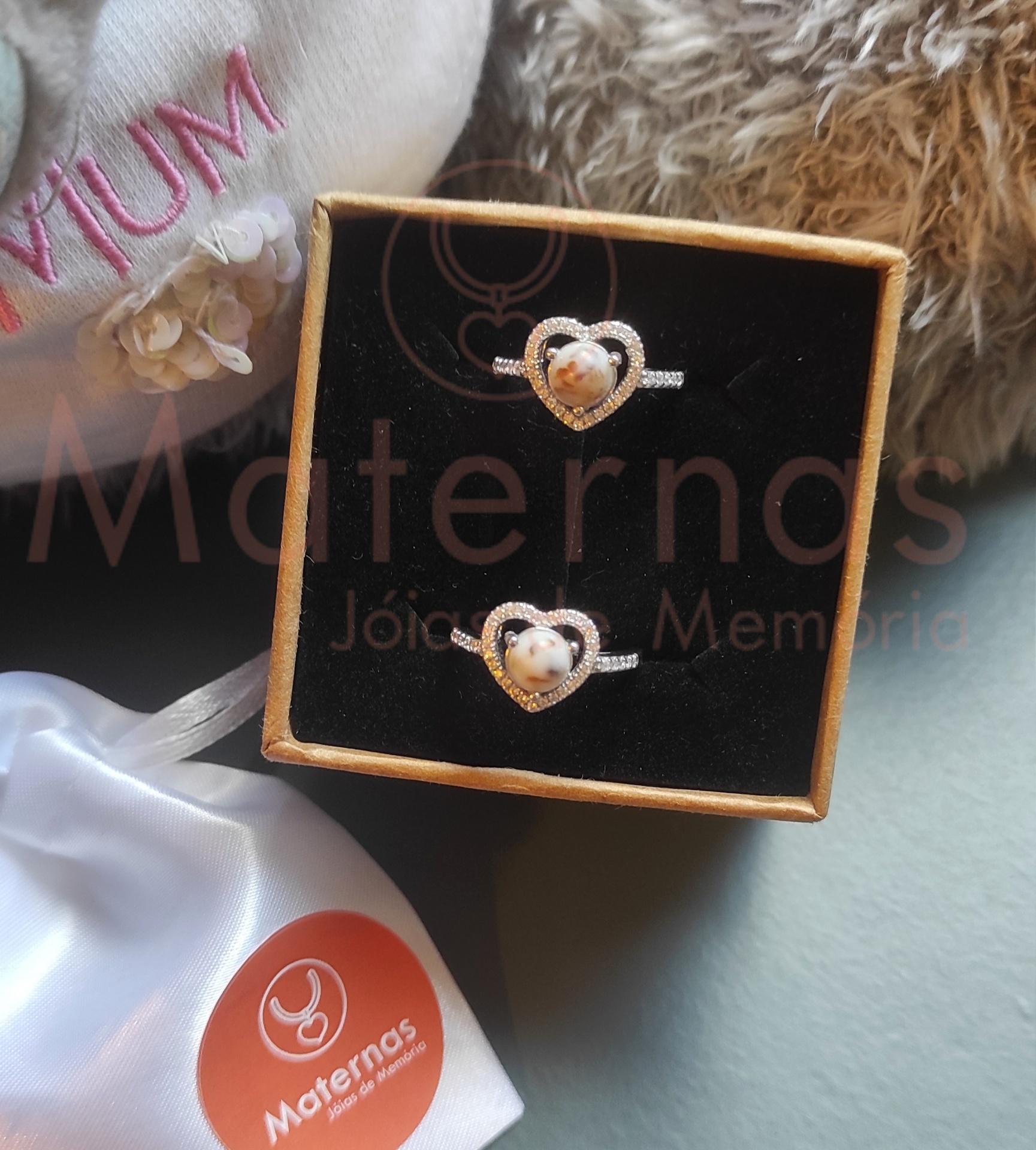 Anel Coração Alto Dois anéis com corações prateados numa caixa preta, saco branco com etiqueta orange Maternas e peluche cinza claro com bordado rosa