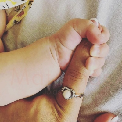 Mão de bebé segurando dedo de adulto com anel prateado em forma de coração.