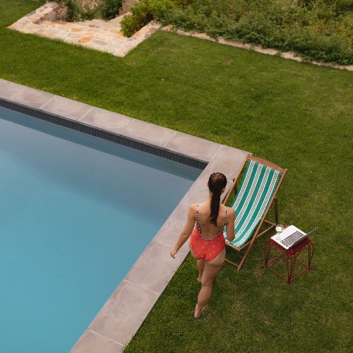 Pessoa com fato de banho vermelho ao lado de piscina, cadeira de praia às riscas e mesa vermelha com portátil e chávena
