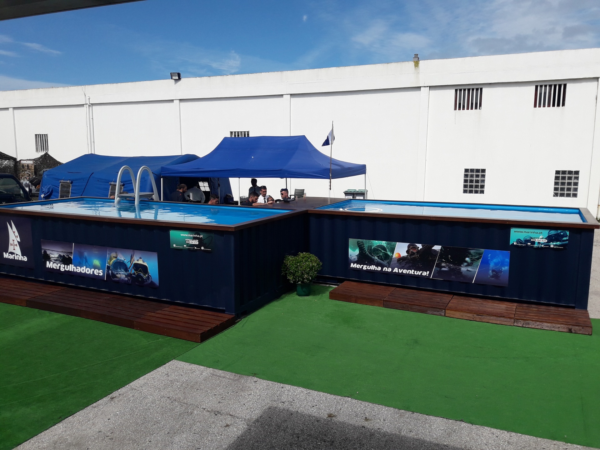 Piscina elevada azul com banners, escada metálica e tendas azuis ao ar livre