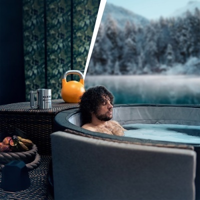 Homem relaxando num jacuzzi redondo cinzento com garrafa, kettlebell e frutas numa mesa de vime ao lado, cenário de floresta e lago gelado ao fundo