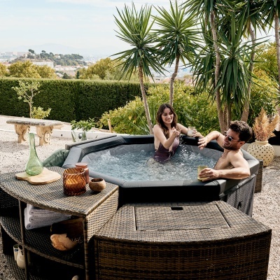 Banheira de hidromassagem cinza escuro ao ar livre com dois adultos sentados, rodeada por mobiliário de vime castanho e plantas tropicais.