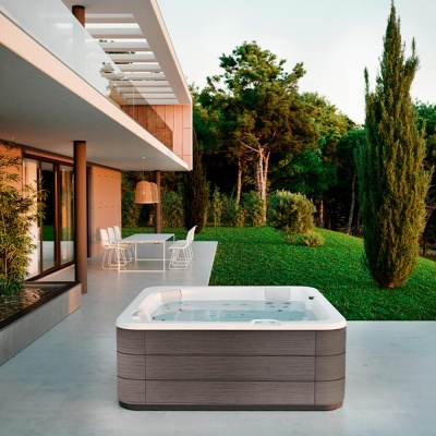 Banheira de hidromassagem exterior quadrada com acabamento em madeira clara e interior branco numa esplanada junto a jardim