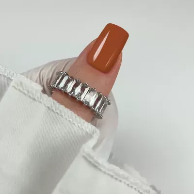 Anel prateado com pedras transparentes em dedo com unha castanho alaranjada e luva branca.