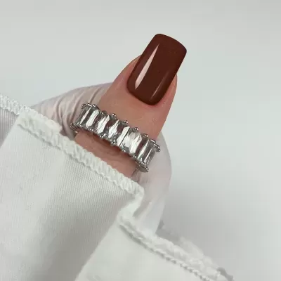 Anel prateado com pedras retangulares em dedo com unha castanha e tecido branco ao fundo