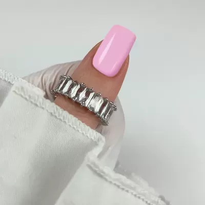 Anel prateado com pedras retangulares num dedo com unha rosa claro e tecido branco.