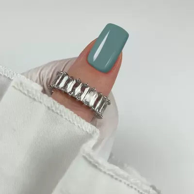 Anel prata com pedras retangulares transparentes em dedo com unha azul acinzentada e manga branca.