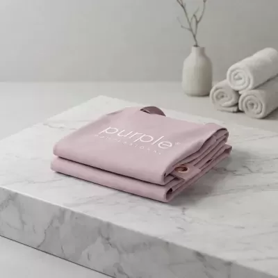 Toalha rosa claro dobrada com letras brancas em superfície de mármore com toalhas enroladas e vaso ao fundo.