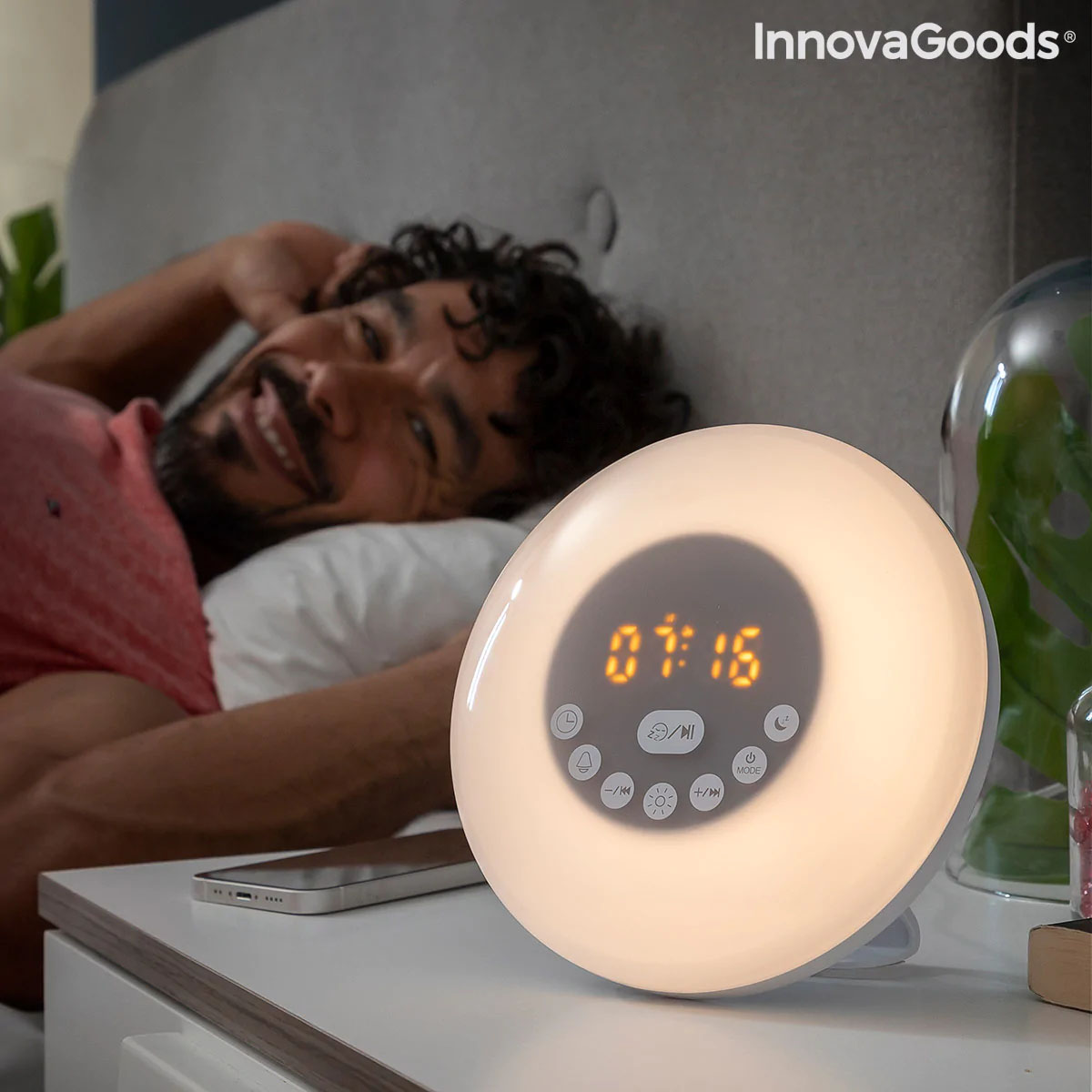 Despertador digital redondo branco com luz e visor LED a mostrar 07:16 numa mesa de cabeceira branca ao lado de smartphone e cama com homem.