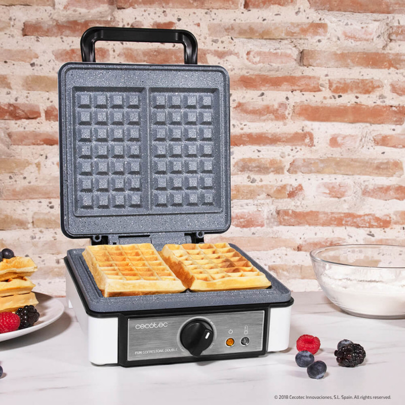 Máquina de waffles Cecotec com waffles na chapa aberta sobre bancada branca e parede de tijolos à vista