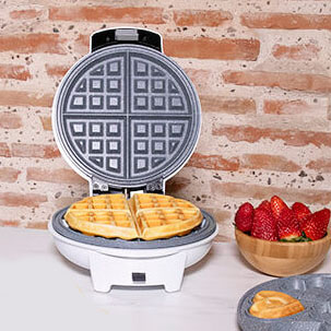 Máquina de waffles branca aberta com waffles dourados e tigela de morangos ao lado numa bancada