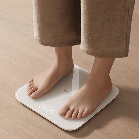 Balança digital branca com mostrador LED e pés descalços sobre chão de madeira