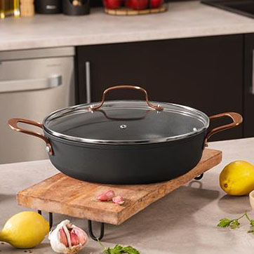 Caçarola preta com tampa de vidro e alças bronze sobre tábua de madeira numa cozinha