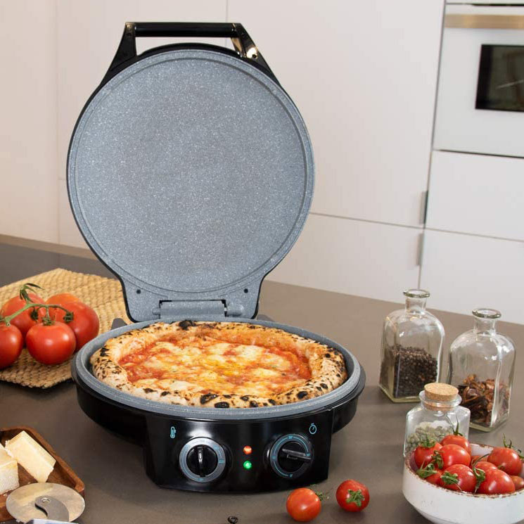 Máquina elétrica para pizza aberta com pizza dentro, tomates, queijo e especiarias ao redor numa cozinha moderna