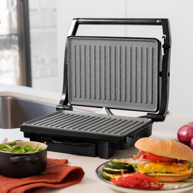 Grill elétrico preto com placas de grelha cinzentas numa cozinha moderna com alimentos ao redor