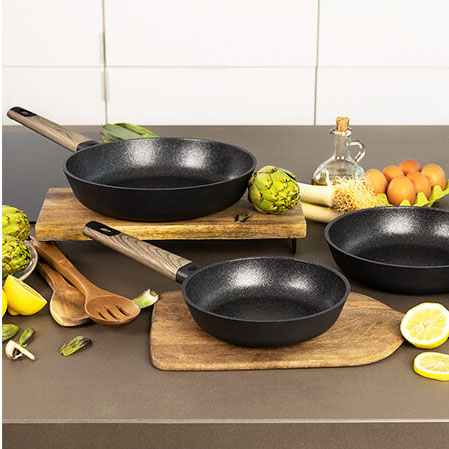 Três frigideiras pretas com cabos de madeira em bancada com ingredientes variados