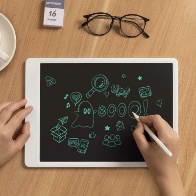Tablet branco com desenhos verdes, caneta digital, óculos pretos e calendário numa mesa de madeira clara.