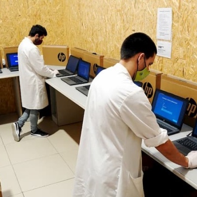 Duas pessoas testando laptops HP numa sala com paredes de madeira OSB e caixas empilhadas