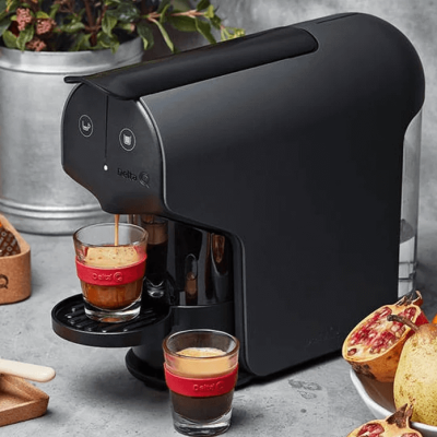 Máquina de café expresso preta com duas chávenas pequenas e frutas partidas ao lado