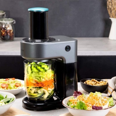 Robot de cozinha a triturar legumes numa taça de vidro, com várias tigelas de salada à volta