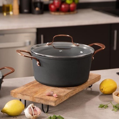 Caçarola preta com tampa de vidro e pega metálica em cobre sobre tábua de madeira na cozinha.