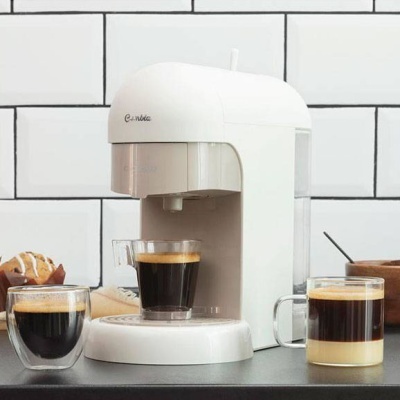 Máquina de café branca Cambia com três chávenas de vidro contendo café, numa bancada cinza com azulejos brancos ao fundo