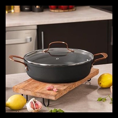Caçarola preta com tampa de vidro e alças bronze sobre tábua de madeira numa cozinha