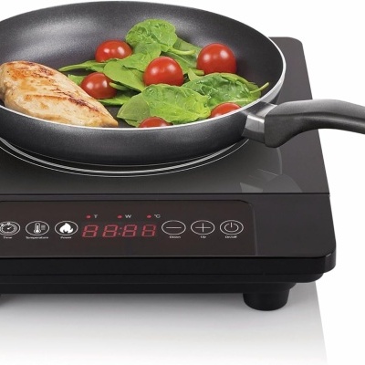 Fogão elétrico preto com frigideira contendo frango, folhas verdes e tomates-cereja