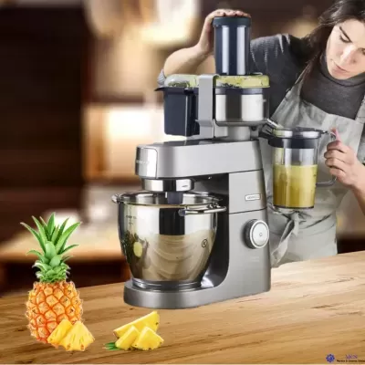 Robot de cozinha metálico prateado com tigela e copo de líquido amarelo, pedaços de ananás e pessoa a usar.