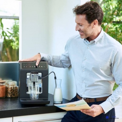 Homem usando máquina de café automática preta num balcão com jarro de leite e frascos de grãos