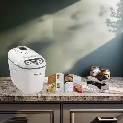Máquina de pão Moulinex branca com painel digital numa bancada com catálogo aberto e potes