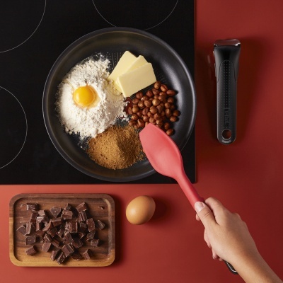 Panela preta com ingredientes para cozinhar e utensílios numa superfície vermelha perto de fogão preto