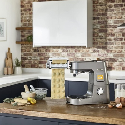 Máquina cinza para fazer massa fresca numa bancada de cozinha com ingredientes e parede de tijolos