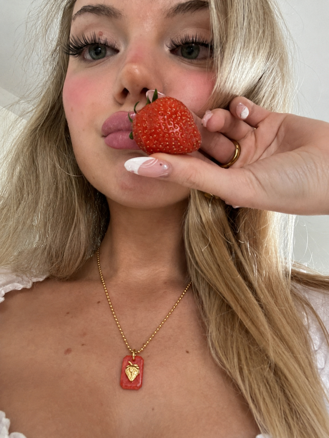 Mulher loira segura morango junto aos lábios e usa colar com pendente vermelho e dourado em forma de morango