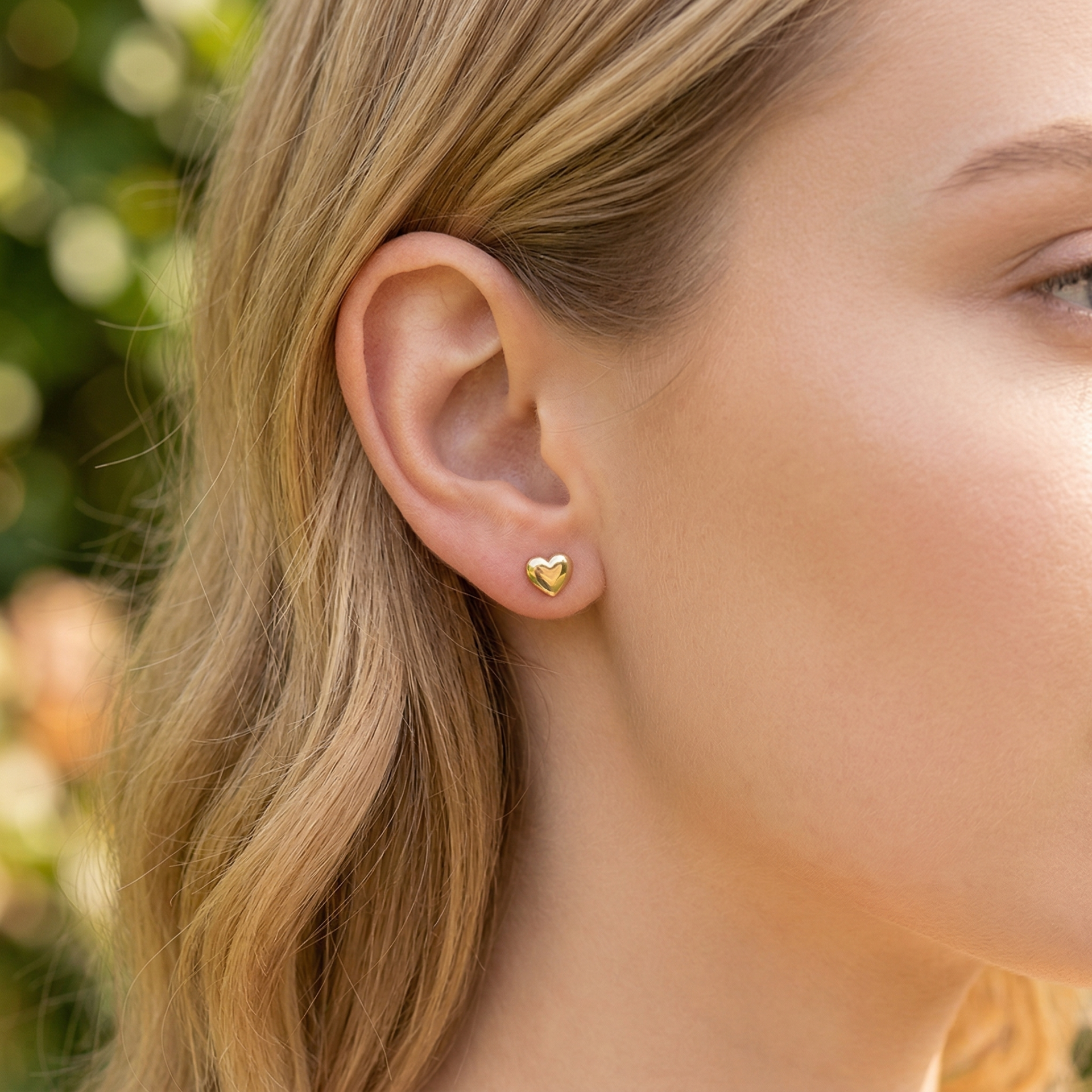 Brinco pequeno coração dourado em ouvido feminino com cabelo louro e ondulado.