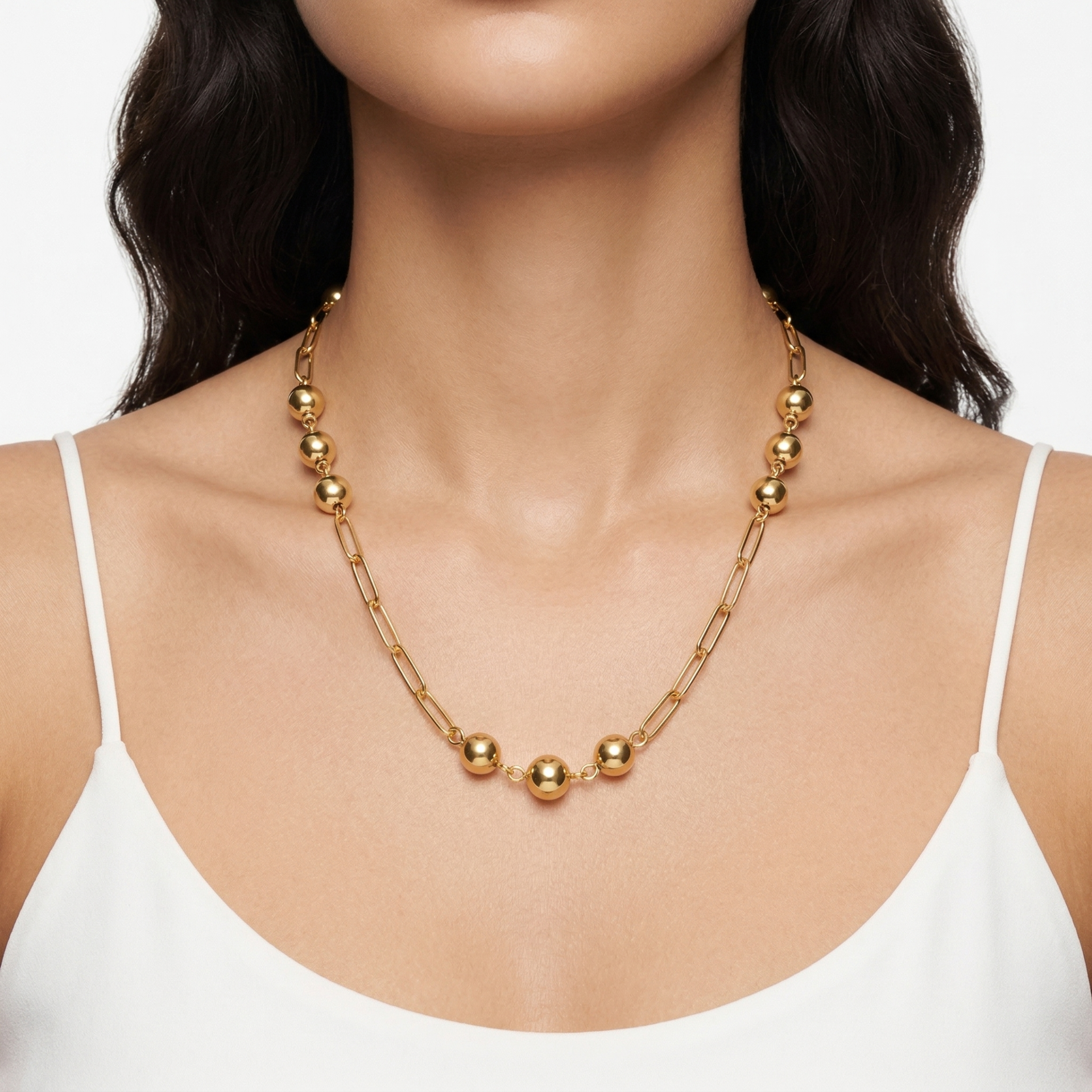 Colar dourado com esferas metálicas e elos, usado ao pescoço por mulher com vestido branco.