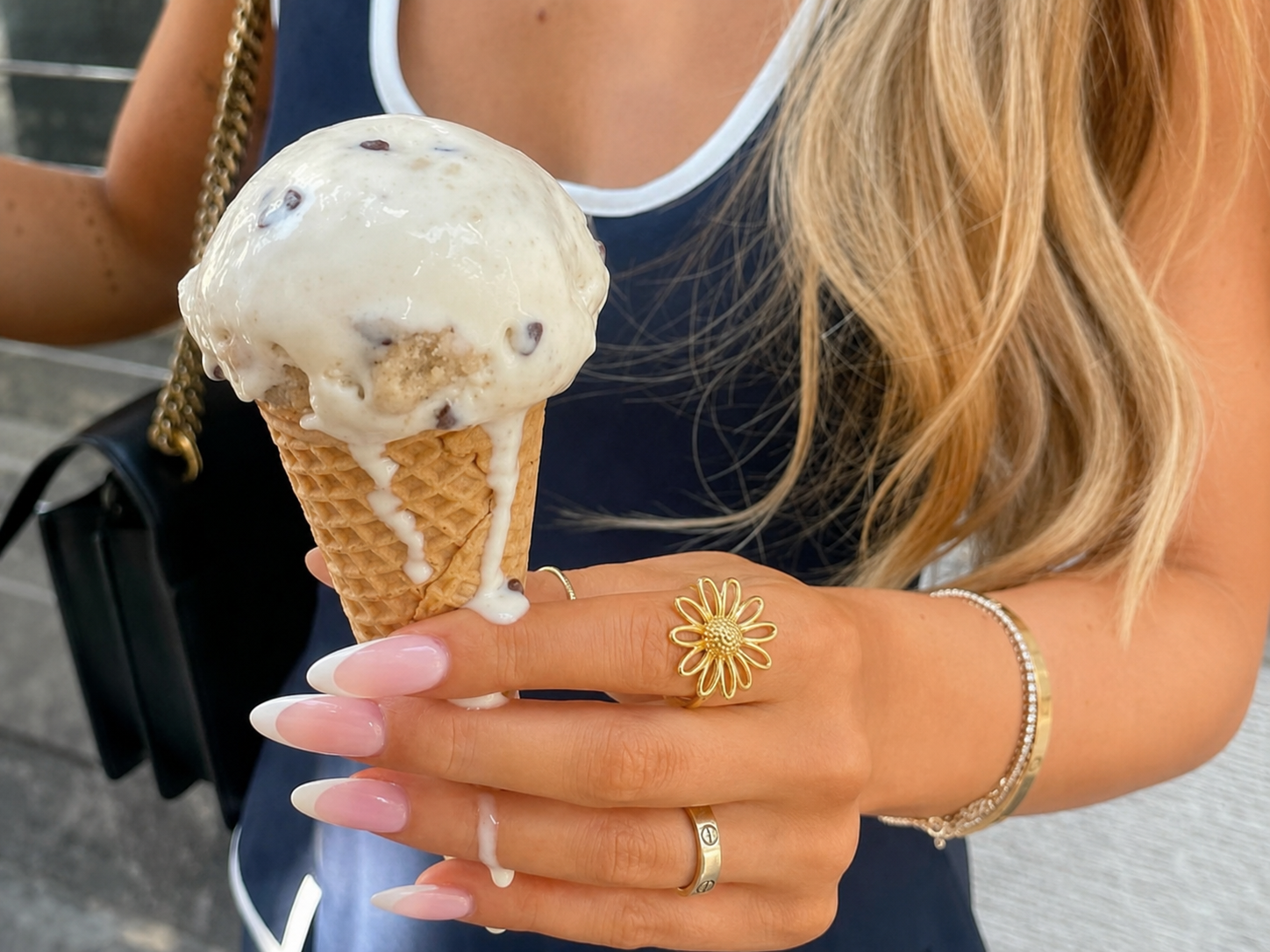 Gelado creme com pedaços de chocolate em cone de waffle, mão feminina com unhas manicuras francesas e joias, fundo com mala preta e roupa azul.