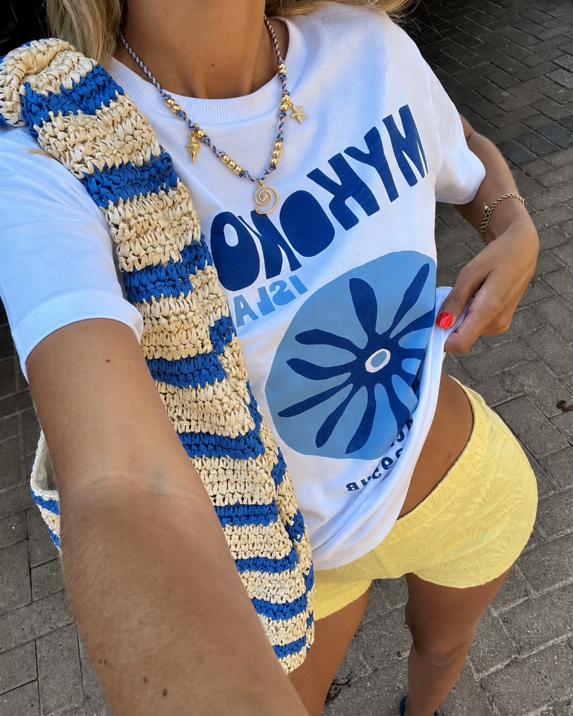 T-shirt branca com estampado azul, shorts amarelos, bolsa de malha azul e bege, colar com contas douradas e pendentes do mar